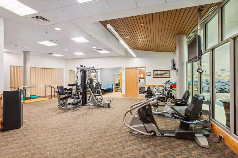 A spacious fitness room in a senior living facility featuring various exercise equipment including stationary bikes, weight machines, and parallel bars. The room has carpeted flooring, large windows on one side, and a wooden ceiling accent. There are televisions mounted on the walls and a door leading to another room.