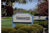 A landscaped entrance sign reading "Homewood" set among trimmed shrubs with trees and a road in the background.