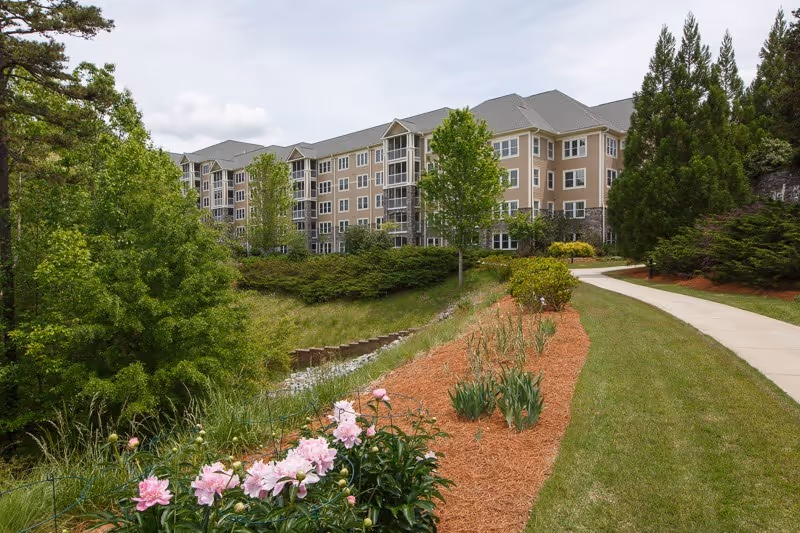 Landscaped walkway and flower beds leading to a multi-story senior living building set among trees.
