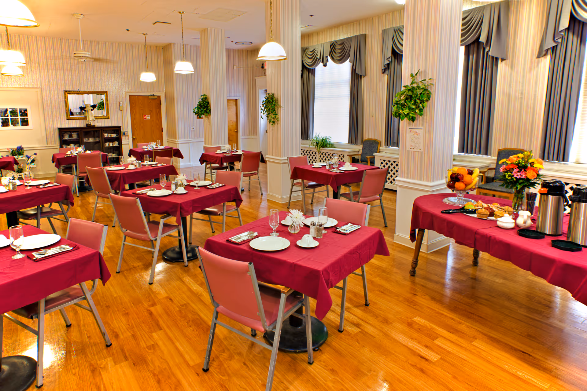 A dining room with multiple tables covered in red tablecloths, each set with plates, glasses, and silverware. Pink chairs surround the tables. The room has wooden floors, large windows with draped curtains, and several hanging pendant lights. A side table holds a fruit bowl, flowers, and coffee dispensers. The walls are decorated with plants and framed pictures.
