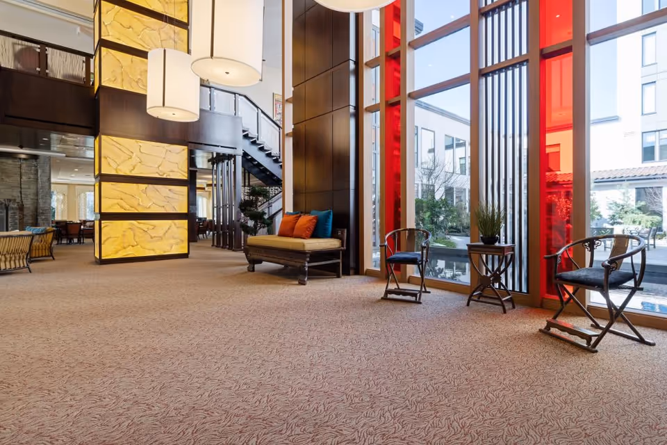 Spacious and modern interior lounge area with large floor-to-ceiling windows featuring red accents, a cushioned bench with colorful pillows, two wooden chairs, a small table with a plant, and a staircase in the background. The room has warm lighting and a patterned carpet.