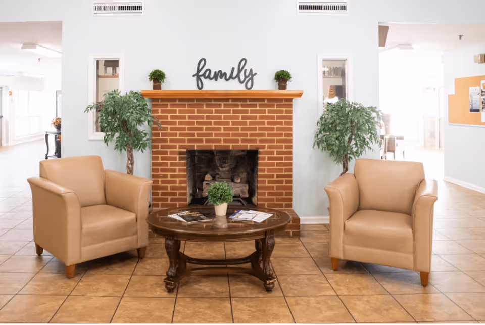 A cozy seating area with two beige armchairs facing a wooden coffee table that holds a small potted plant and some magazines. Behind the table is a red brick fireplace with the word 'family' displayed above it. Two green potted plants flank the fireplace, and the room has light-colored tiled flooring and walls painted in a soft pastel shade.