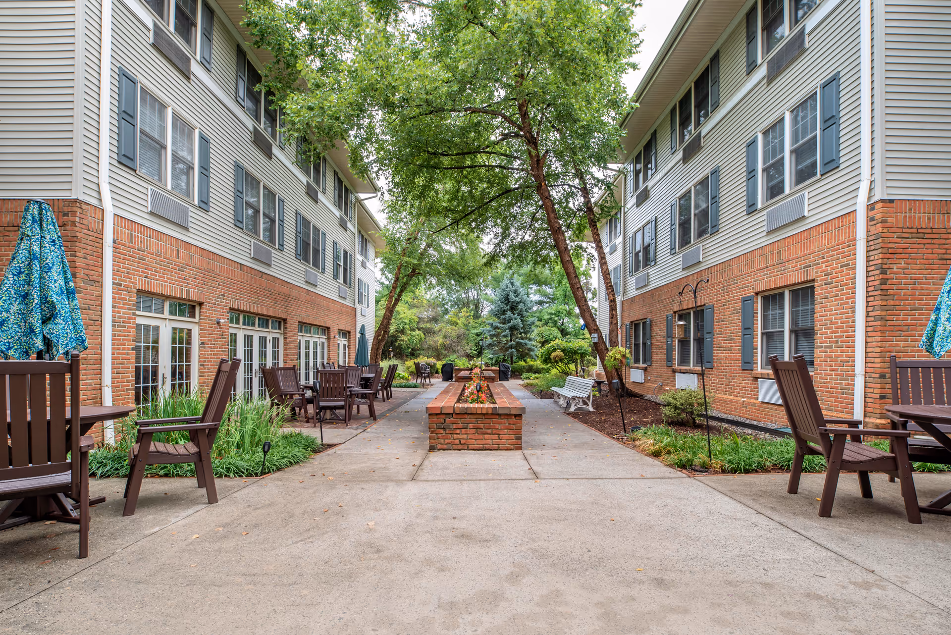 Outdoor courtyard area between two brick and siding buildings with patio tables, chairs, umbrellas, a central brick planter, trees, and greenery.