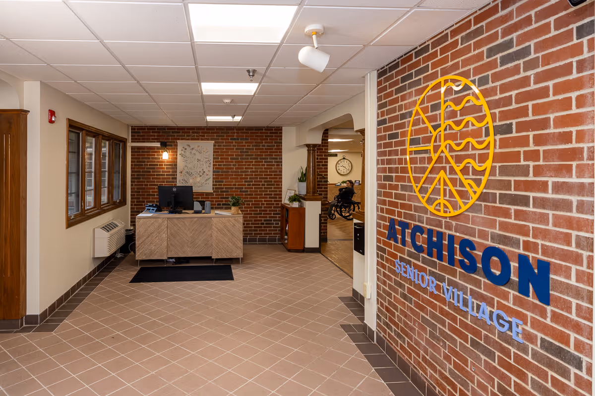 Interior view of the entrance area at Atchison Senior Village featuring a brick wall with the facility's logo and name, a reception desk with a computer, and a hallway leading to other rooms. The floor is tiled and the ceiling has recessed lighting.