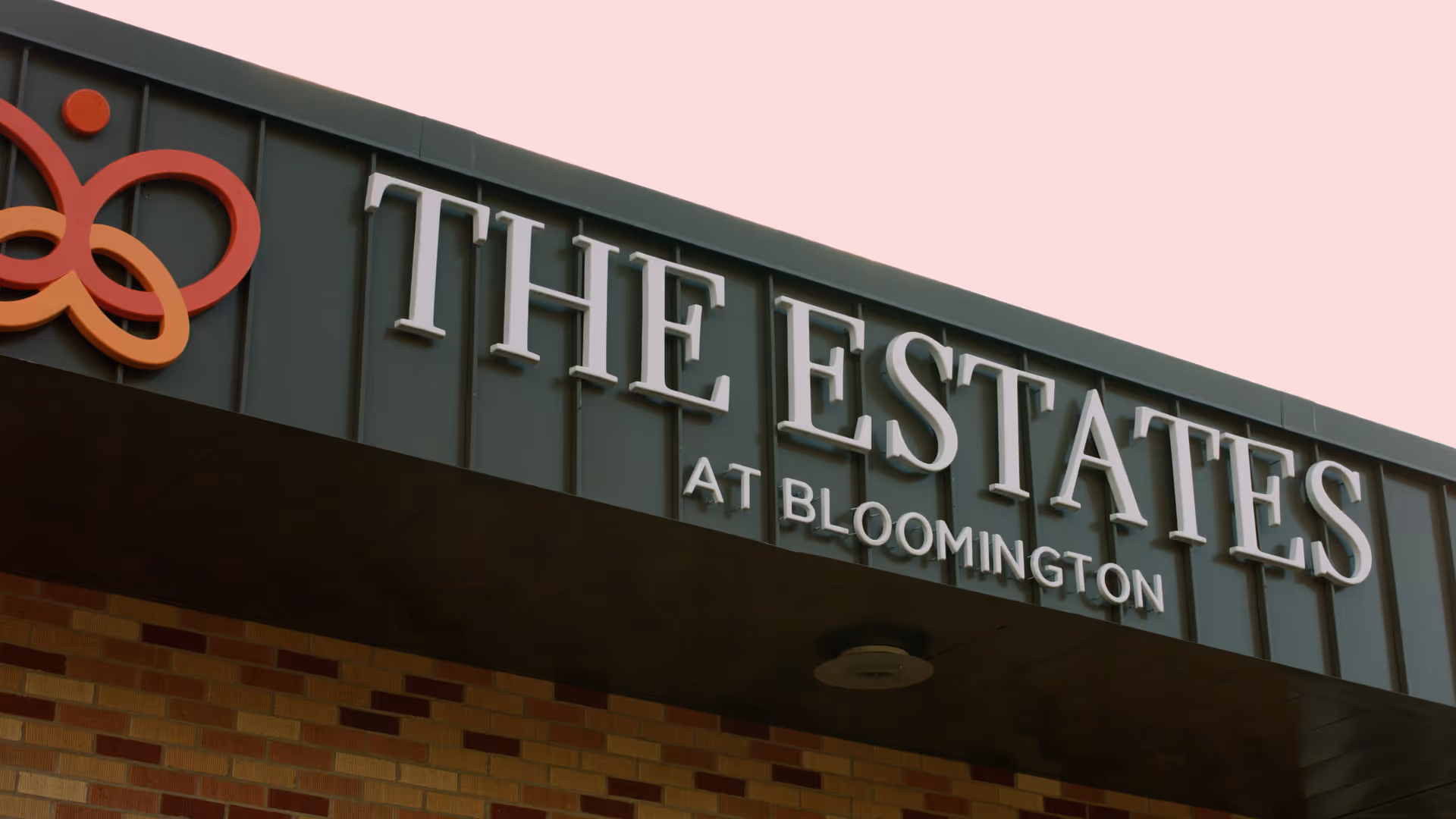 Close-up view of the exterior sign for The Estates at Bloomington on a building with a brick facade and a dark green panel background.