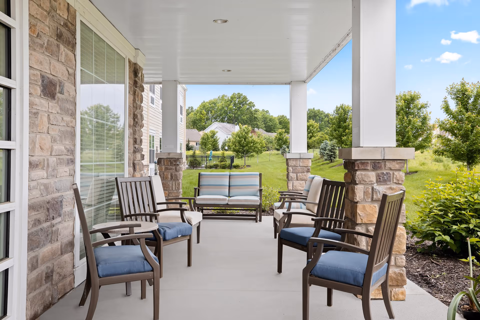 Covered outdoor patio area with stone pillars and several cushioned chairs arranged facing each other. The patio overlooks a grassy yard with trees and neighboring houses in the background under a blue sky with some clouds.