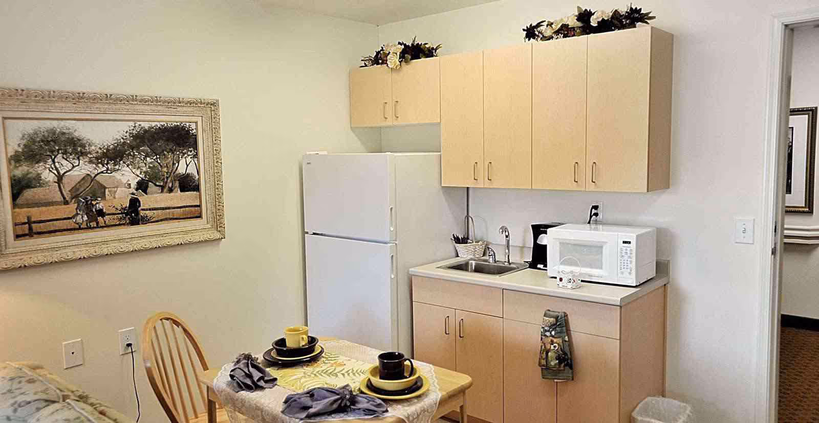 A small kitchenette area with light wood cabinets, a white refrigerator, a microwave, a coffee maker, and a sink. In front of the kitchenette is a small dining table set for two with yellow and black dishes, cups, and napkins. A framed painting of a rural scene with trees and people hangs on the wall to the left. The room has light-colored walls and carpeted flooring.