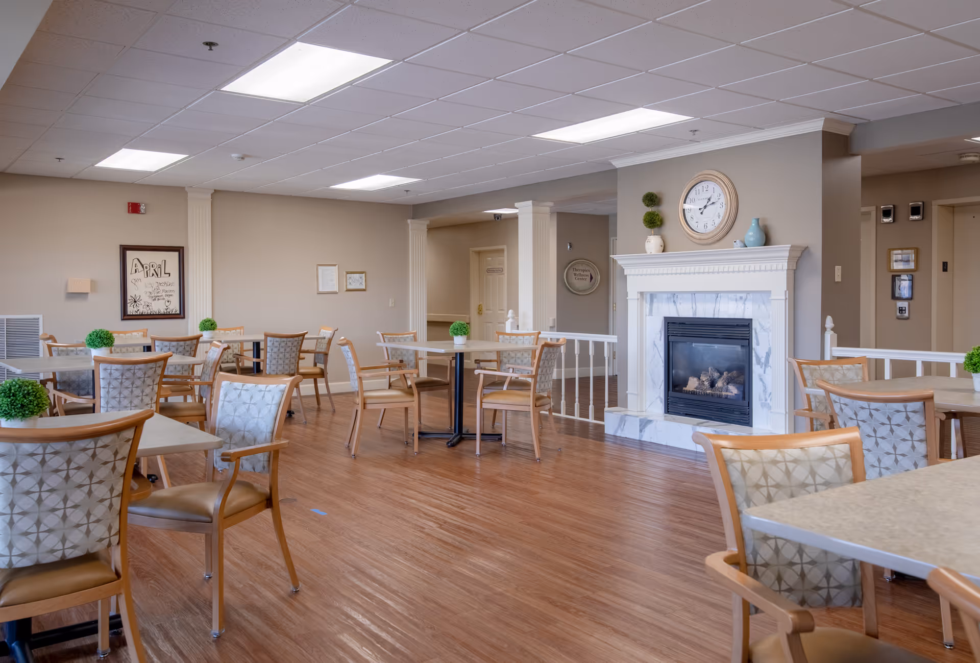 A bright and spacious dining area with multiple tables and chairs arranged neatly. The room features a wooden floor, a white fireplace with decorative items and a clock above it, and beige walls. Small green plants are placed on some tables, and there is a framed artwork on one wall.