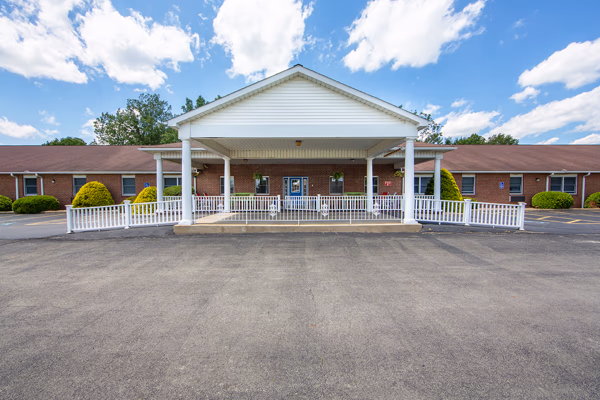 Front exterior view of a single-story brick building with a covered entrance supported by white columns. The building has a brown roof and is surrounded by neatly trimmed bushes and a paved parking area. The sky is partly cloudy with blue patches visible.