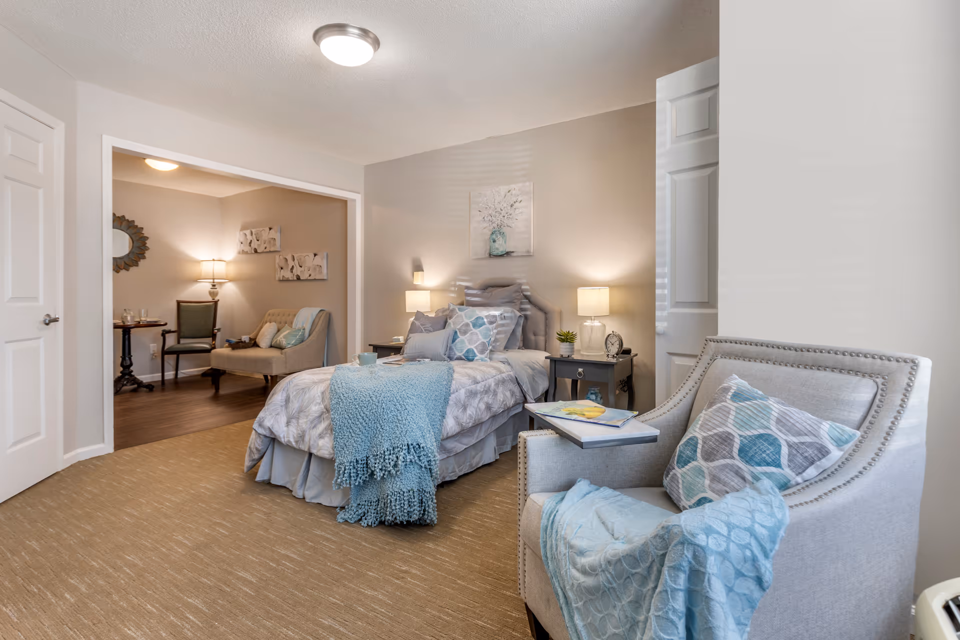 A cozy bedroom in a senior living facility featuring a single bed with multiple pillows and a blue throw blanket. Next to the bed are two nightstands with lamps, a small plant, and an alarm clock. A comfortable armchair with a patterned pillow and a blue throw is positioned in the foreground. In the background, there is a small sitting area with a loveseat, a chair, a side table, and a lamp. The walls are decorated with artwork and a mirror, and the room has soft, warm lighting.