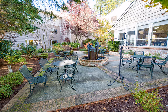 Outdoor patio area at The Gardens at Marysville featuring several black metal tables and chairs arranged around a central brick water fountain, surrounded by trees and shrubs with a building in the background.