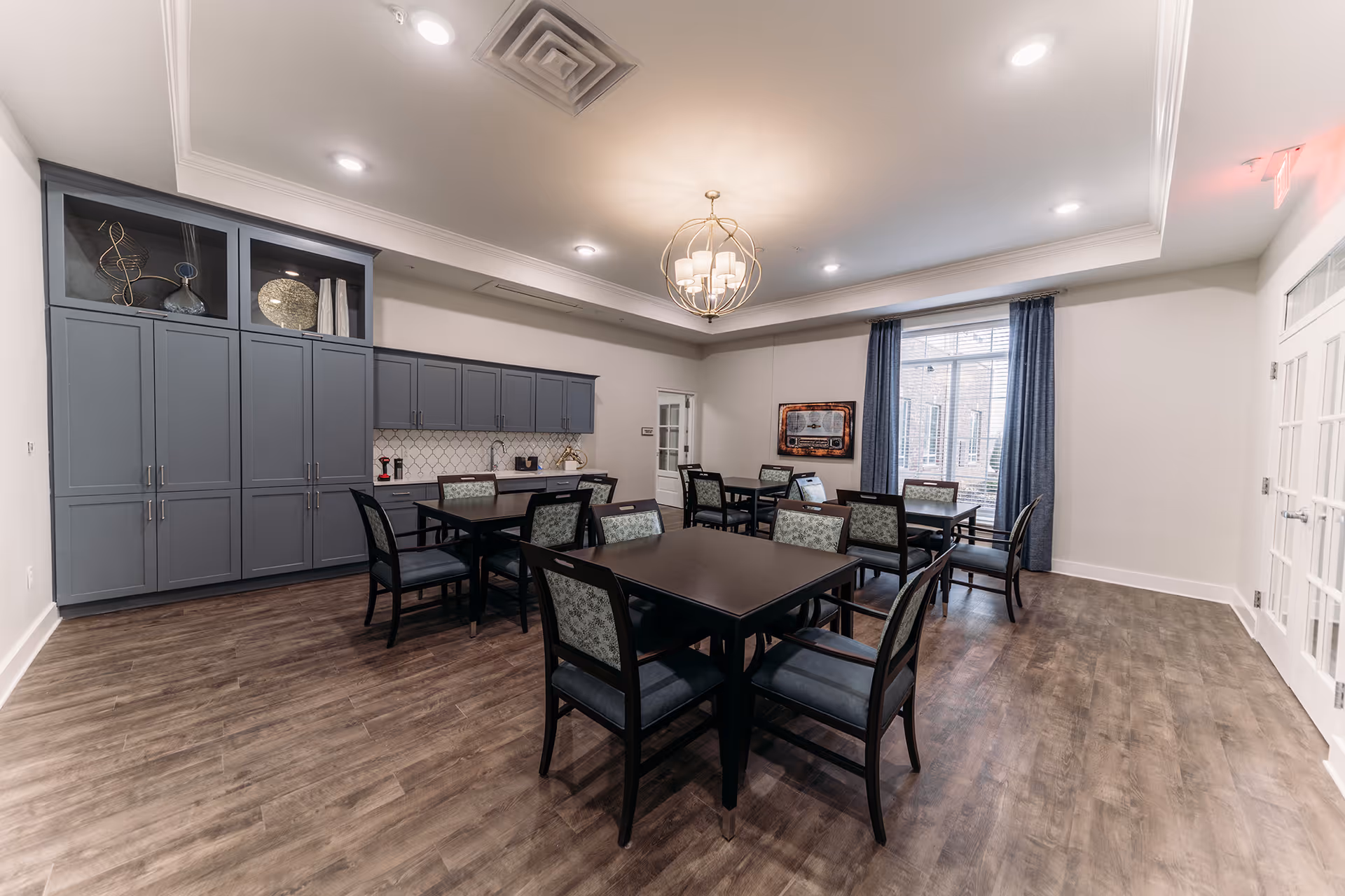 A well-lit dining room with several dark wood tables and chairs with patterned upholstery. The room features wood flooring, gray cabinetry along one wall with decorative items on top, a modern chandelier hanging from the ceiling, and a large window with blue curtains letting in natural light.