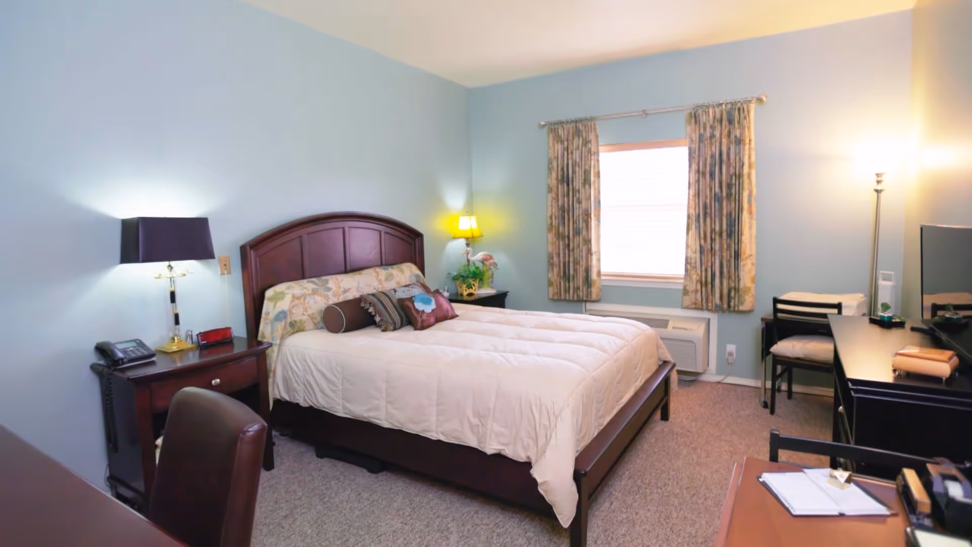 A cozy bedroom with light blue walls, a bed with a wooden headboard and beige bedding adorned with decorative pillows. There are two nightstands with lamps on either side of the bed. A window with floral curtains is on the far wall. A desk with a chair and a floor lamp are positioned near the window, and a TV is on a dresser to the right.