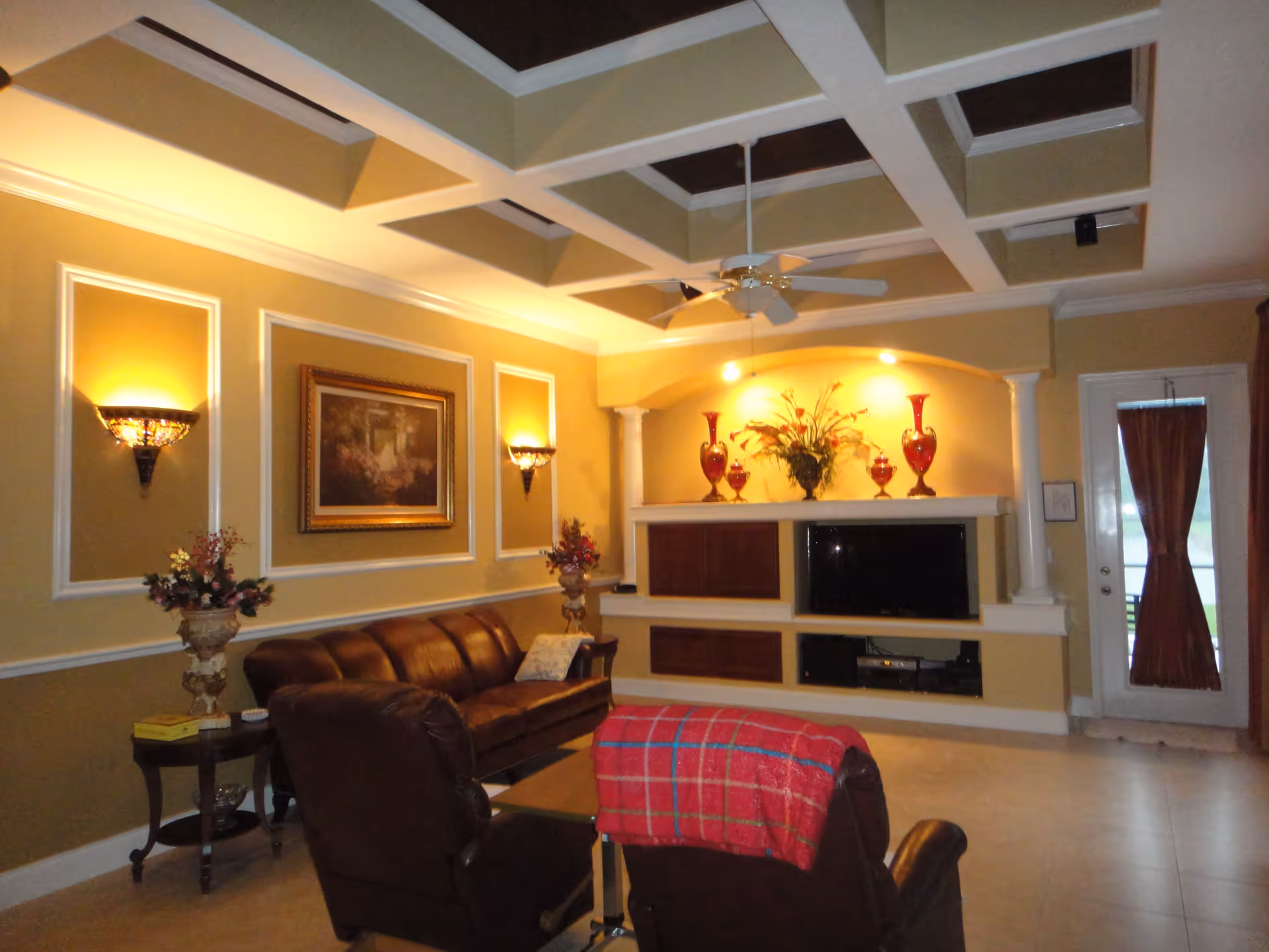 A cozy living room with a brown leather sofa and two armchairs, one draped with a red plaid blanket. The walls are painted yellow with white trim and decorated with a framed painting and wall sconces. A built-in entertainment center holds a flat-screen TV and decorative red vases with flowers. A ceiling fan hangs from a coffered ceiling, and a door with a red curtain leads outside.