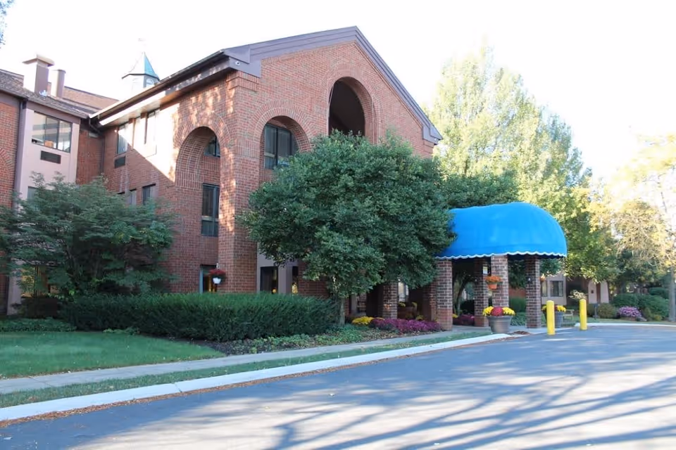 Brick senior living facility entrance with blue awning and landscaped shrubs.