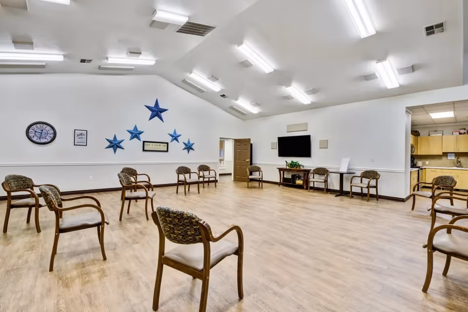 A spacious room with wooden flooring and white walls, arranged with several wooden chairs spaced apart in a circular pattern. The walls feature blue star decorations, a clock, and framed signs. There is a large flat-screen TV mounted on one wall above a small table with a plant. The room is brightly lit with multiple ceiling lights, and a doorway leads to a kitchen area with wooden cabinets.