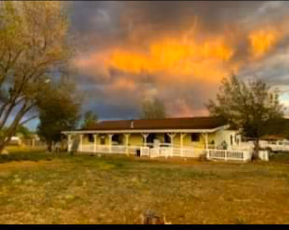 A single-story building with a brown roof and a white porch railing, surrounded by trees and grass under a dramatic sky with orange and gray clouds at sunset.