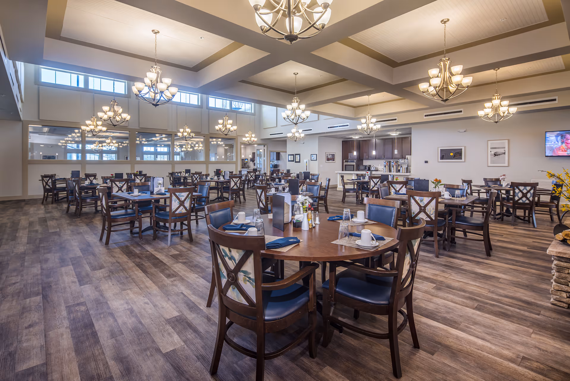 Spacious dining room with multiple wooden tables and chairs arranged neatly. Each table is set with cups, glasses, napkins, and small flower arrangements. The room features large windows near the ceiling, multiple chandeliers hanging from a coffered ceiling, and a kitchen area in the background with dark cabinetry and stainless steel appliances.