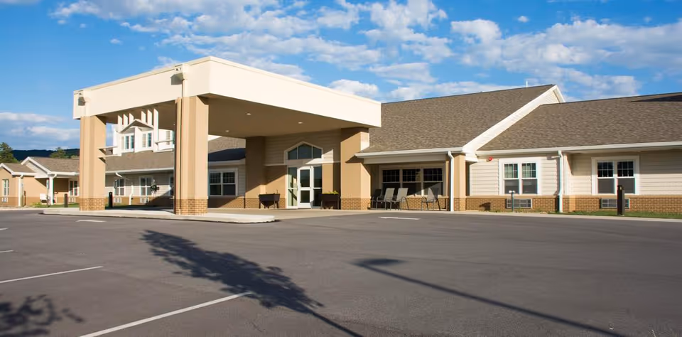 Front entrance and parking area of a single-story senior living building with a covered porte-cochère under a blue sky.