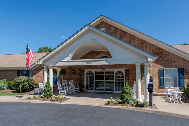 Front exterior view of a single-story brick building with a covered entrance supported by white columns. There are rocking chairs on the porch, an American flag on the left side, and a small garden with shrubs and flowers near the entrance. The sky is clear and blue.