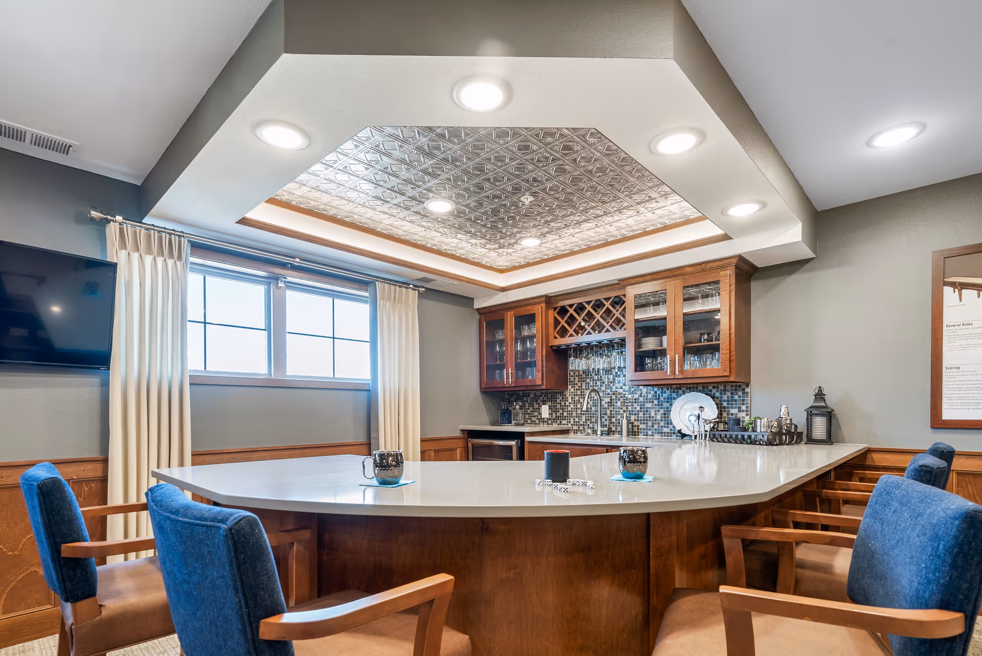 A well-lit communal kitchen and bar area with a large curved countertop, upholstered chairs, wood cabinetry, and a decorative ceiling.