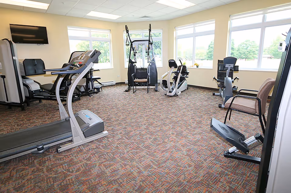 A fitness room in Pleasant View Lutheran Home with various exercise equipment including a treadmill, stationary bikes, and strength training machines. The room has large windows letting in natural light and a wall-mounted TV.