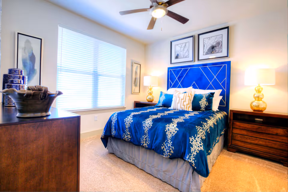 A bright bedroom featuring a bed with a blue and white patterned comforter and matching pillows, a blue upholstered headboard, two wooden nightstands with gold lamps, a ceiling fan with light, a window with blinds, and decorative artwork on the walls.
