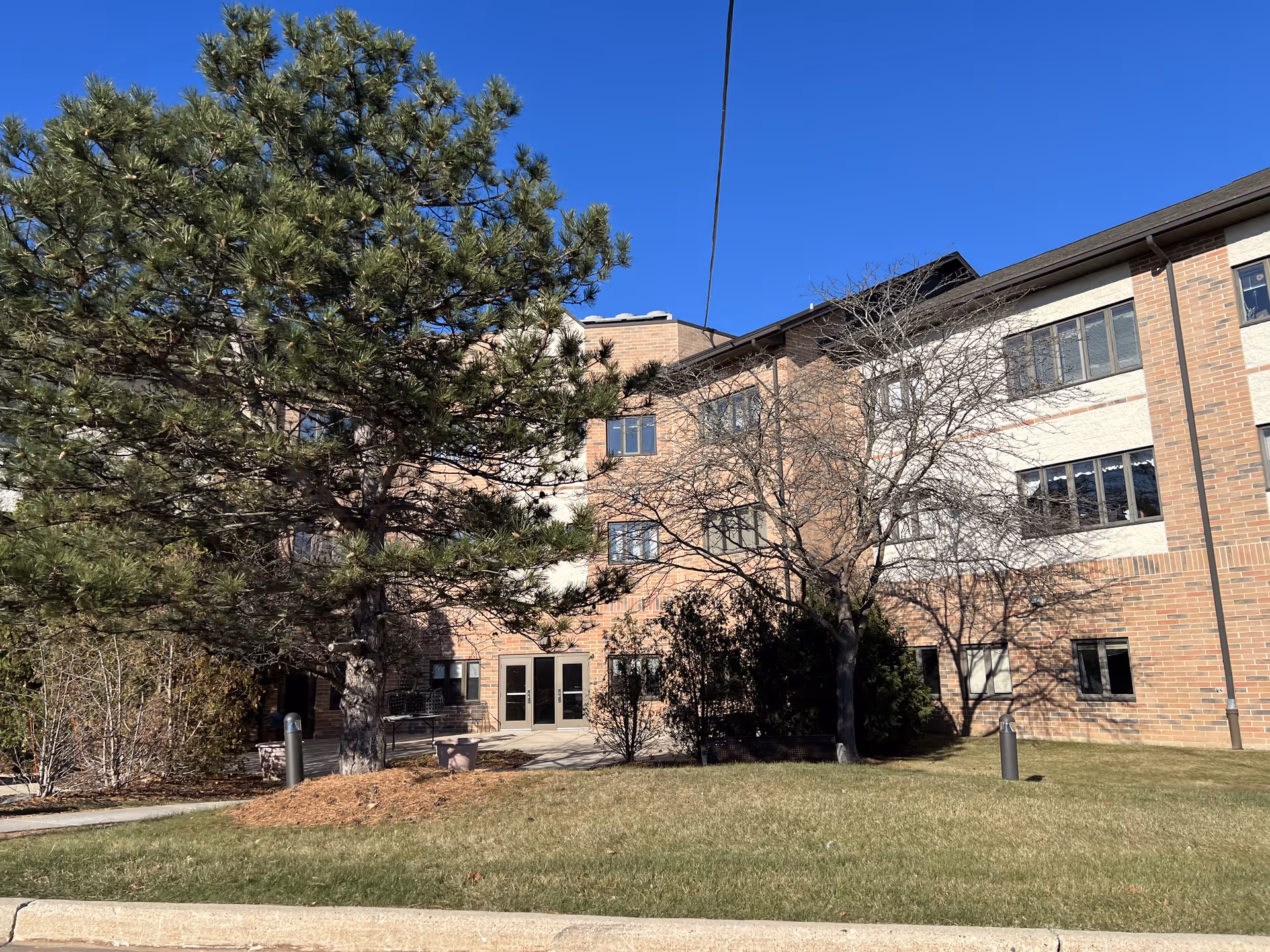Exterior view of a multi-story brick building with several windows, surrounded by trees and a grassy lawn under a clear blue sky.