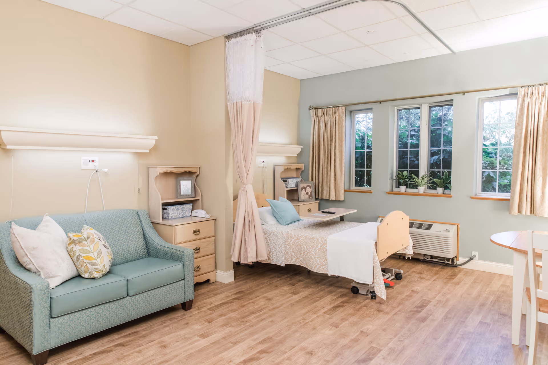 A bright and clean nursing facility room with a hospital bed, a green loveseat with decorative pillows, two small wooden nightstands, a privacy curtain, three windows with beige curtains, and a small round table with chairs. The room has light-colored walls and wood flooring.