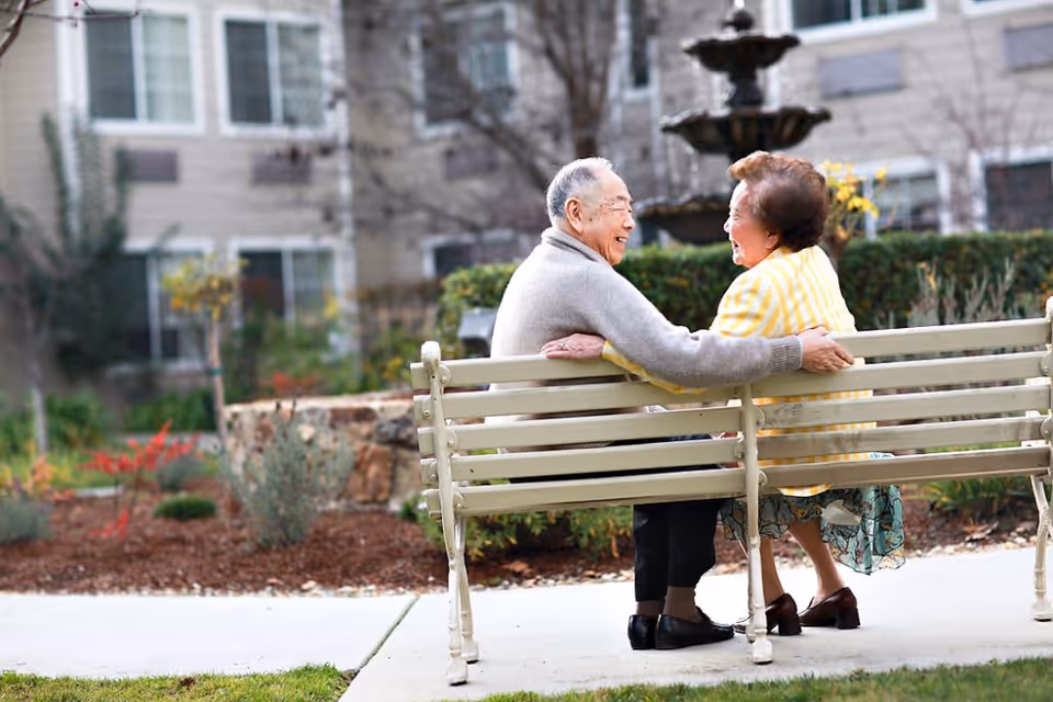 An elderly couple sitting on a bench in a garden area of a senior living facility, smiling and holding each other. There is a multi-tiered fountain and residential building in the background.