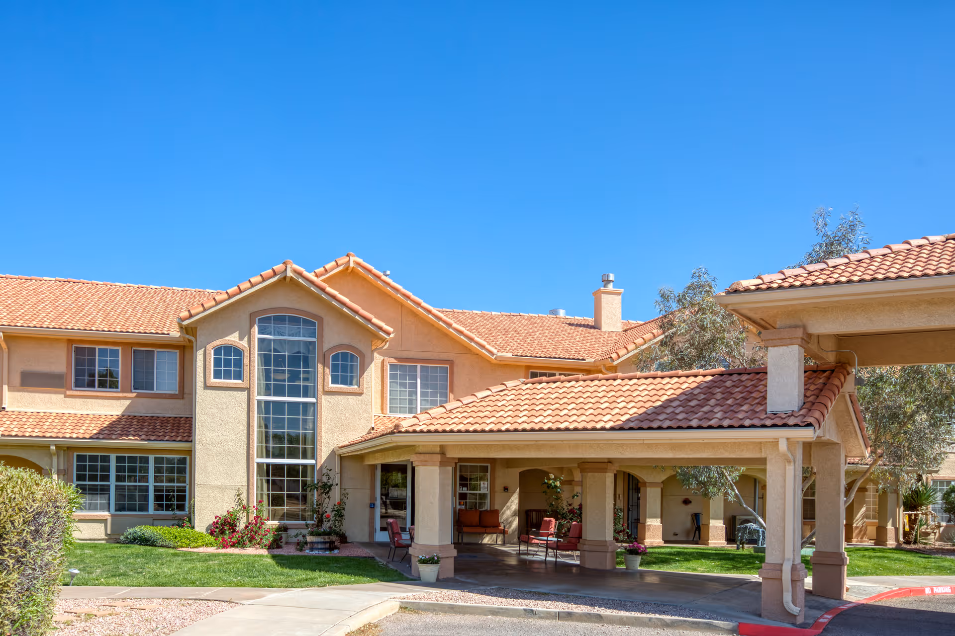 Exterior view of a senior living facility building with beige walls and a red tile roof under a clear blue sky. The entrance area has a covered driveway with seating and potted plants. There are large windows and well-maintained landscaping with green grass and bushes.