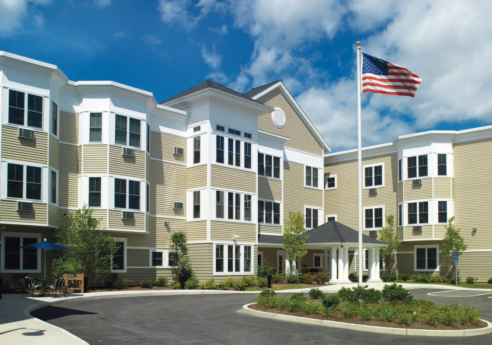 Exterior view of a three-story senior living facility building with beige siding and white trim. The building features multiple large windows and a covered entrance supported by white columns. An American flag is flying on a flagpole in front of the building under a partly cloudy blue sky. There is a circular driveway and landscaped greenery around the entrance.