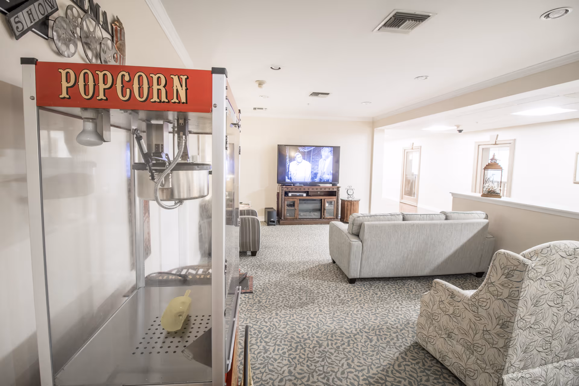 A cozy lobby area featuring a vintage popcorn machine, a gray sofa, and a television displaying a black-and-white film. The room has a patterned carpet and light-colored walls, creating a welcoming atmosphere.