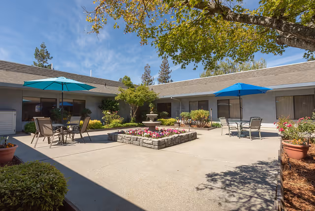 Outdoor courtyard area at Crystal Ridge Care Center featuring a central stone planter with colorful flowers and a small fountain. There are patio tables with blue umbrellas and chairs arranged around the courtyard. The building surrounds the courtyard with windows and greenery, under a clear blue sky.