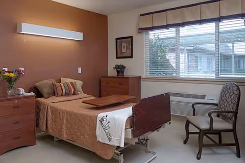 Cozy hospital-style bedroom with a bed, dressers, a window, and a chair.