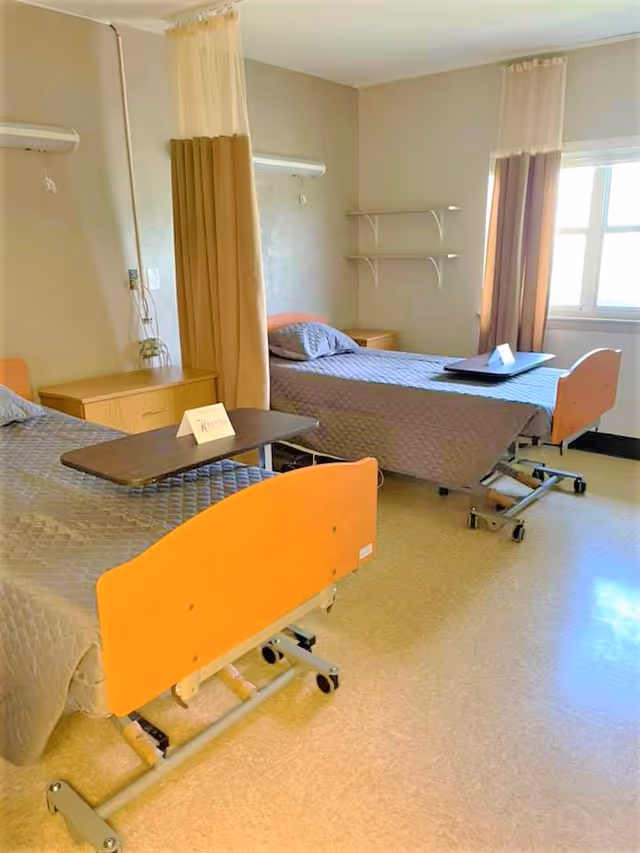 A room with two hospital-style beds separated by a beige privacy curtain. Each bed has a gray quilted blanket and a pillow. There are overbed tables on each bed, one with a small sign that reads 'Kerens'. The room has beige walls, a window with beige curtains, two small wooden nightstands, and two white wall shelves.