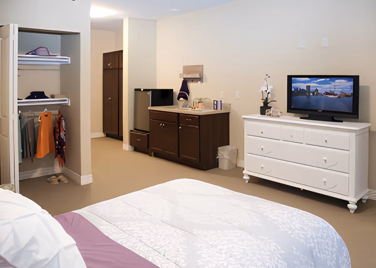 Interior view of a senior living facility bedroom at Danbury Brunswick featuring a bed with white and purple bedding, an open closet with hanging clothes and shelves, a small kitchenette area with a mini fridge and sink, and a white dresser with a flat-screen TV on top. The walls are light-colored and the floor is carpeted.