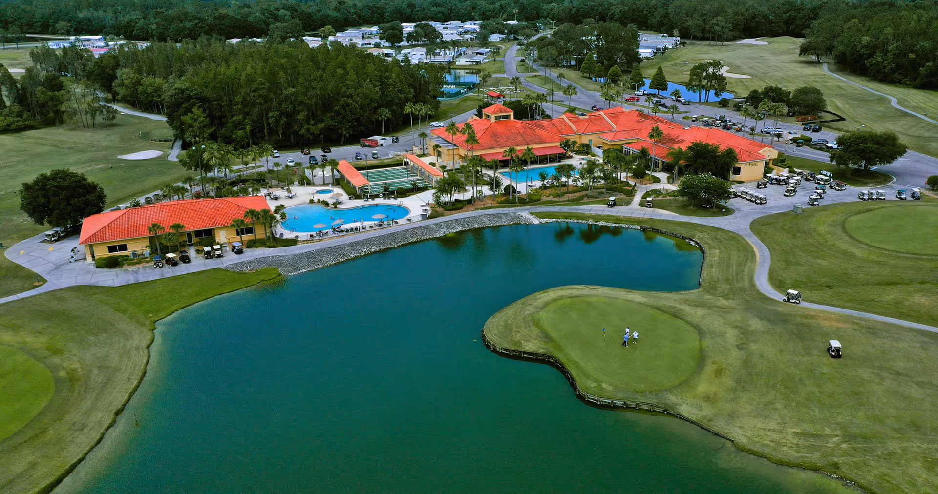 Aerial view of Cypress Lakes Village showing a large pond with a golf green and golf carts on the surrounding golf course. The facility buildings with red roofs, a swimming pool, tennis courts, and parking areas are visible. Trees and residential areas are in the background.