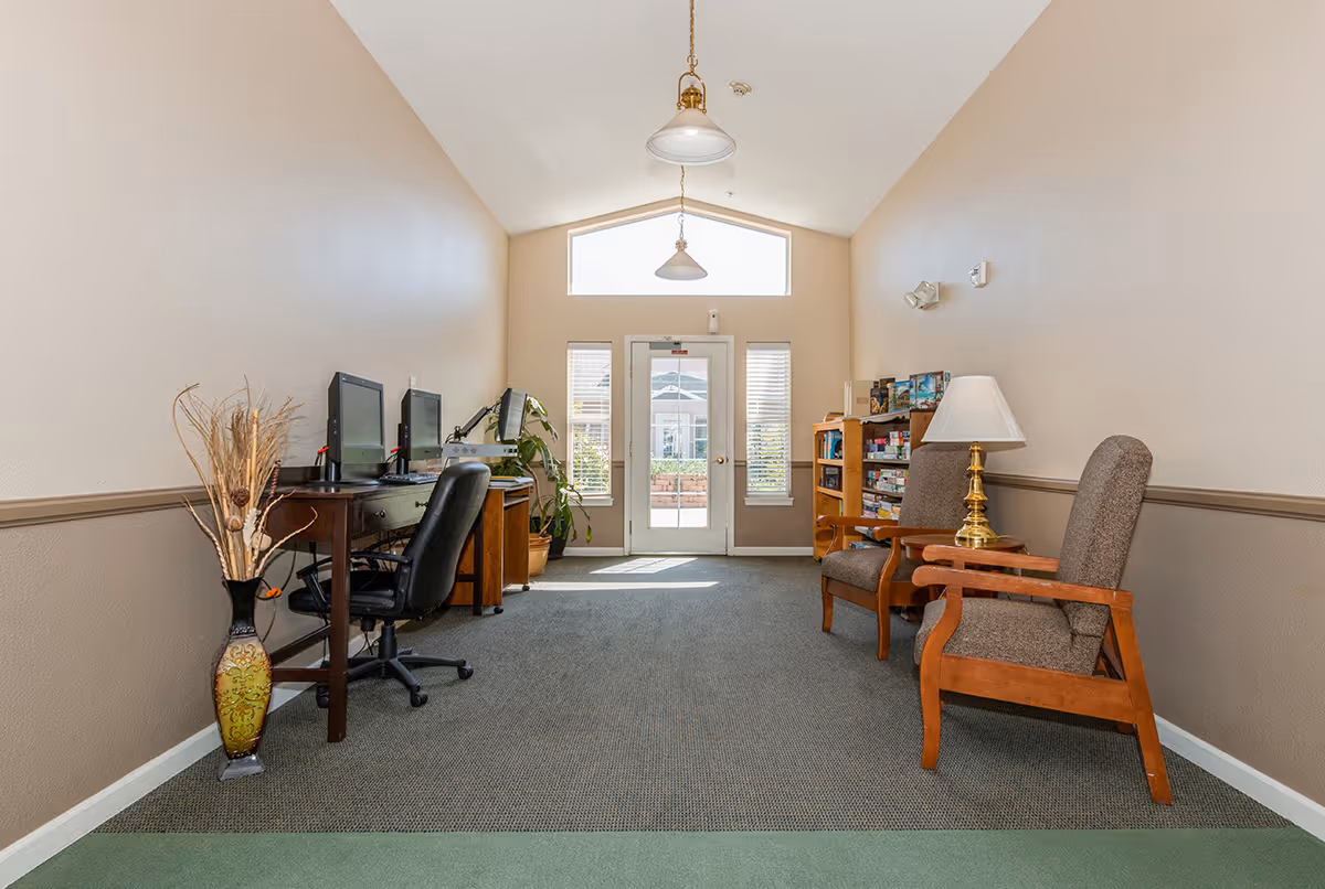 A bright room with beige walls and a high ceiling featuring two hanging pendant lights. On the left side, there is a desk with two computer monitors and a black office chair. A decorative vase with dried plants is placed on the floor next to the desk. On the right side, there are two wooden armchairs with brown cushions and a small wooden side table with a brass lamp. A wooden bookshelf filled with board games and books is also visible. At the far end, there is a glass door flanked by two windows, allowing natural light to enter the room.