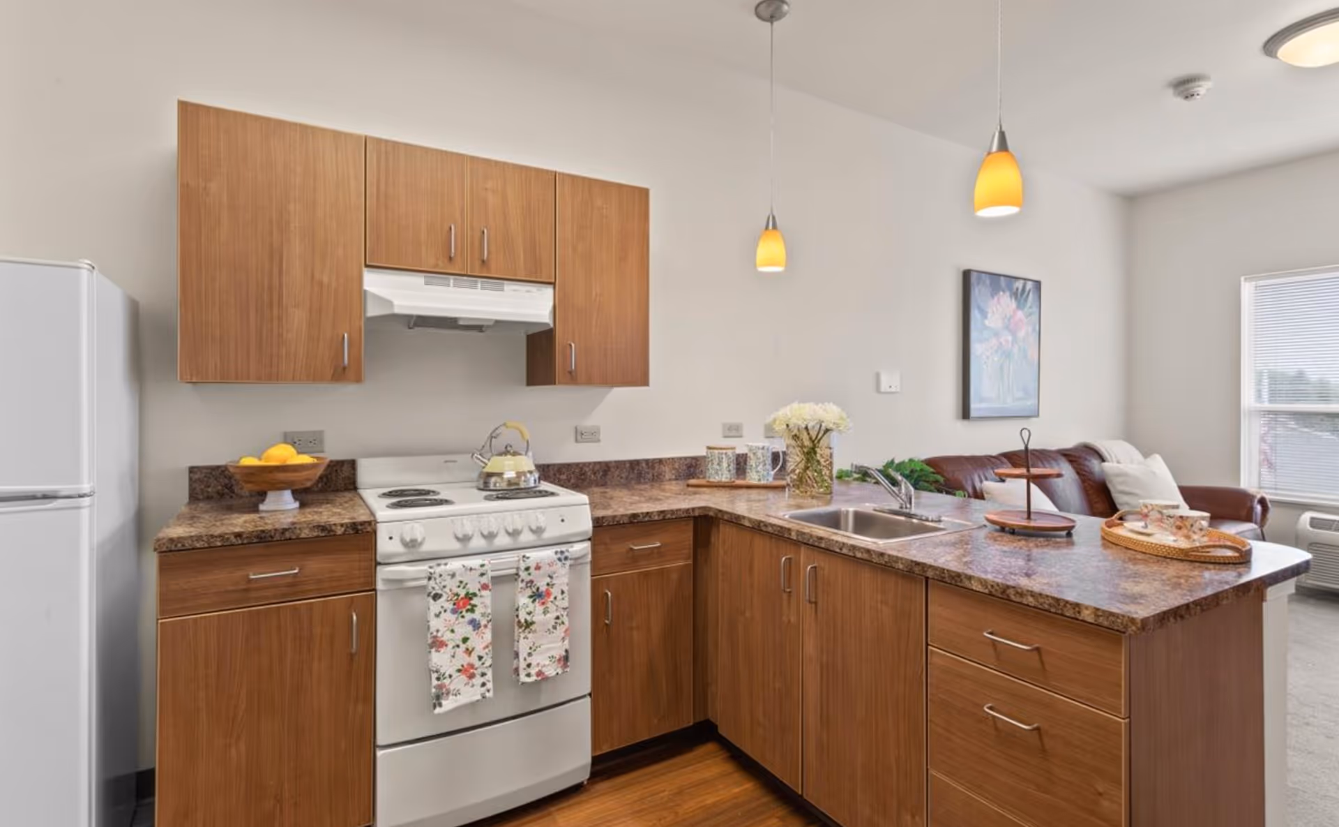 A modern kitchen area with wooden cabinets and a white stove with floral-patterned towels hanging on the oven door. The countertop has a bowl of lemons, a kettle on the stove, a vase with white flowers, and a tiered tray. Two pendant lights hang from the ceiling. In the background, there is a brown leather couch, a framed floral painting on the wall, and a window with blinds letting in natural light.