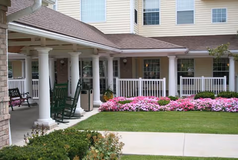 Outdoor patio area of a senior living community with white columns, green rocking chairs, a bench, and a garden bed filled with pink flowers in front of a beige building with multiple windows.