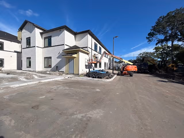 Exterior view of a two-story building under construction with white siding and black roof trim, construction equipment and materials on the ground, and a clear blue sky overhead.