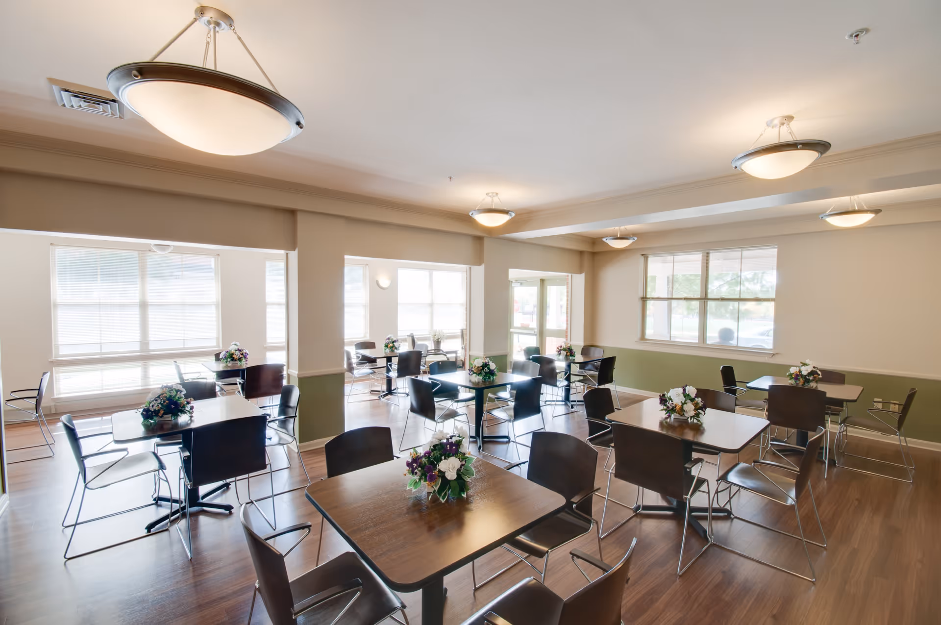 A bright dining room with multiple square wooden tables, each surrounded by four black chairs. Each table has a small floral centerpiece. The room has large windows allowing natural light to fill the space, light-colored walls with a green lower half, and modern ceiling lights.