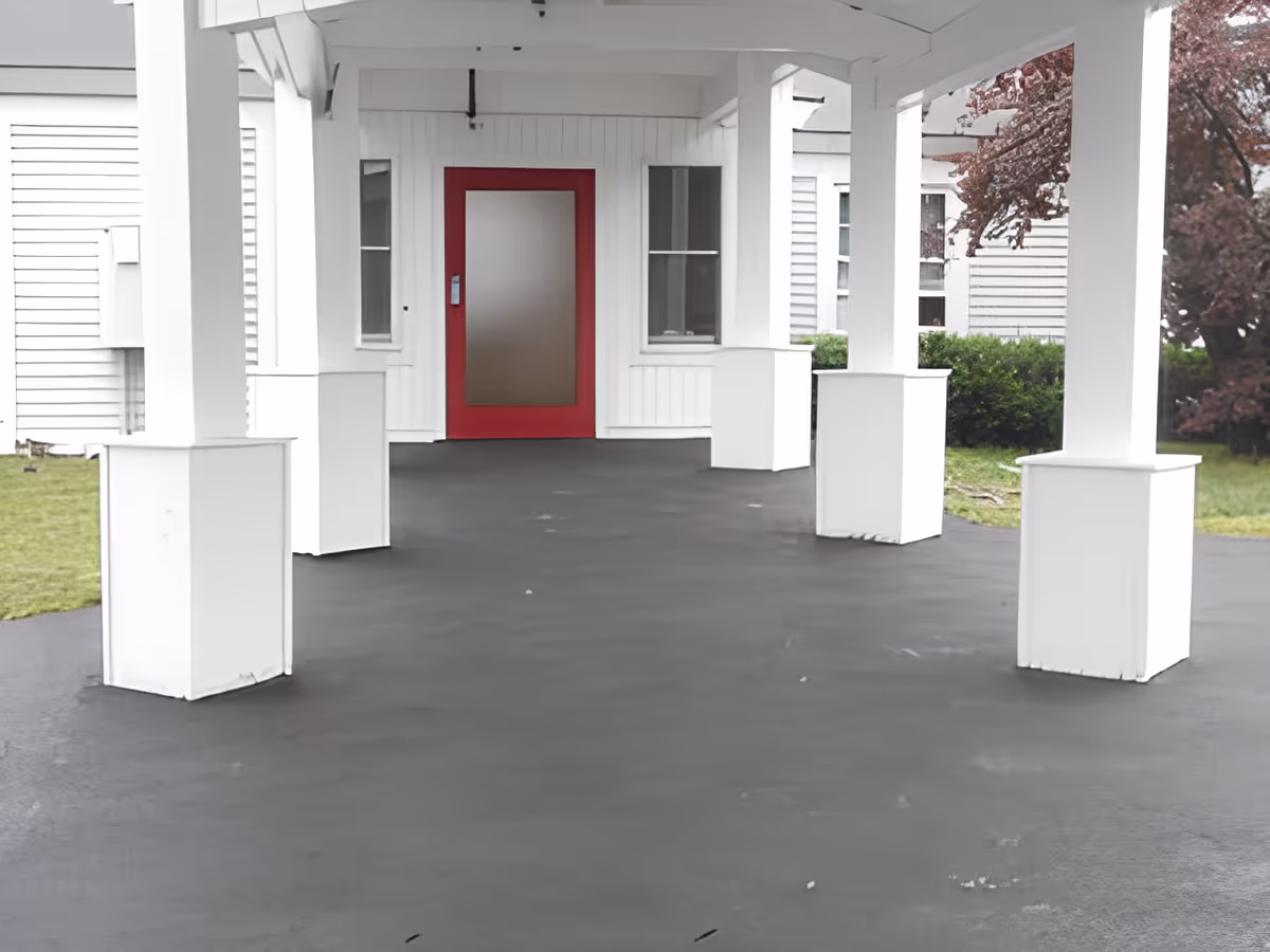Covered entrance area with white pillars and a red door at the end, surrounded by white siding walls and some greenery visible on the sides.