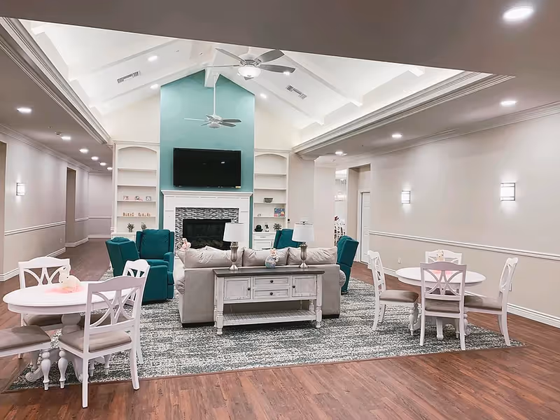 Bright, open living room with teal seating, white tables, and a fireplace with a TV.