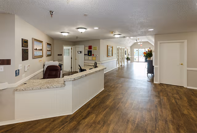 Reception area inside Grayson Court facility with a curved granite countertop desk, office chair, framed pictures on the wall, and a long hallway with wooden flooring leading to glass doors at the far end.