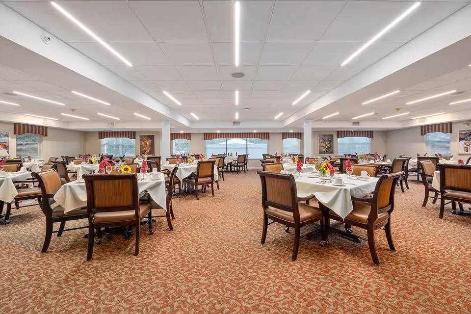 Spacious dining room with multiple round tables set with white tablecloths, chairs, and floral centerpieces under bright ceiling lights.