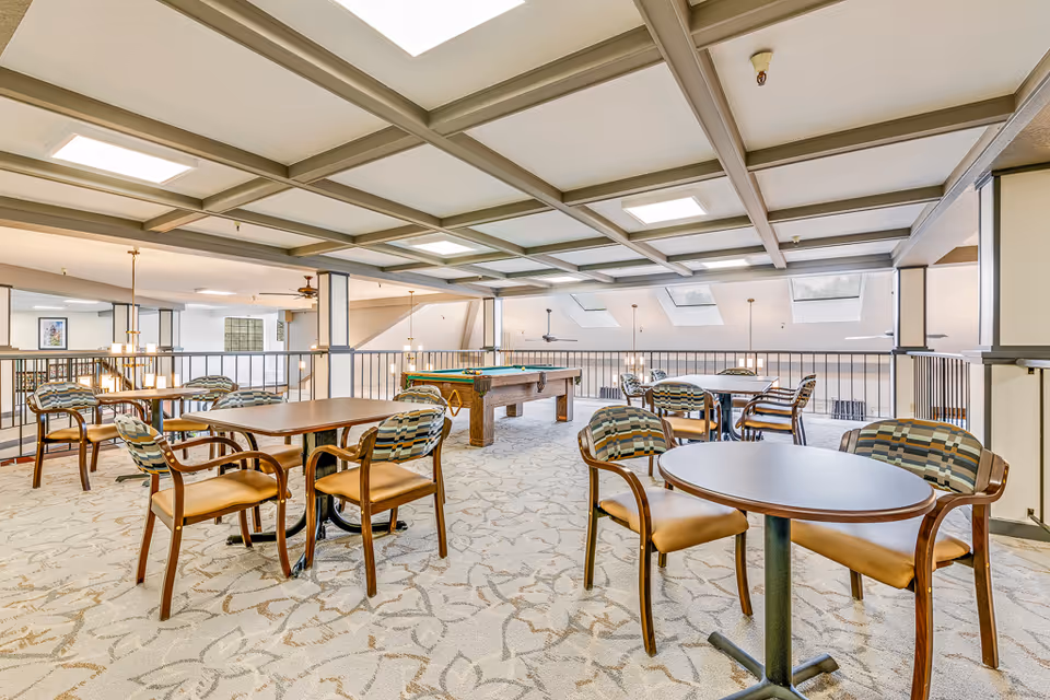 Bright communal lounge with round tables, upholstered chairs, and a pool table overlooking an open atrium.