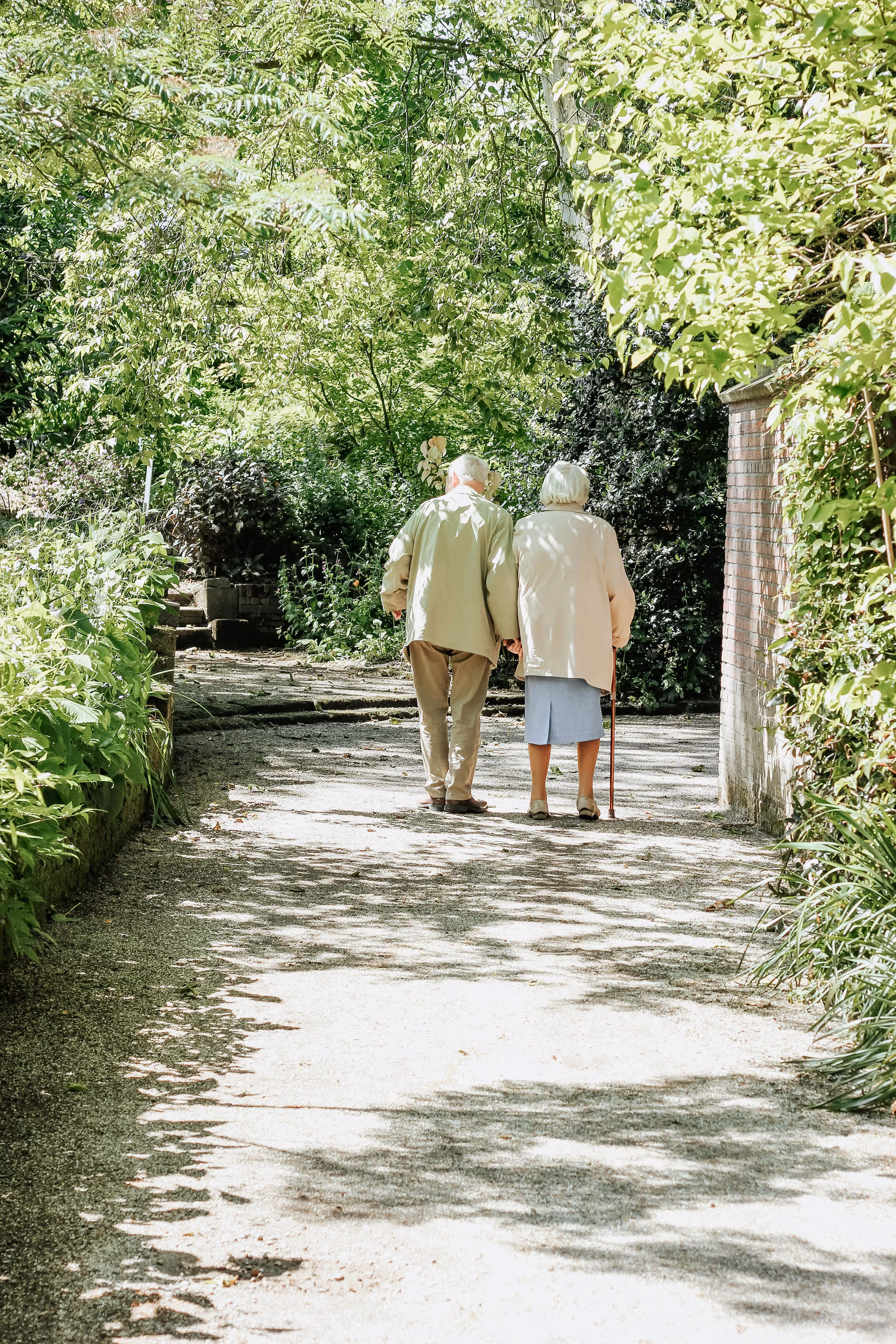 An elderly couple walking together on a shaded garden path surrounded by lush green trees and plants. The woman is using a cane and they are seen from behind.
