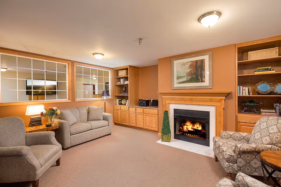 Cozy living room with beige carpet, a lit fireplace with a wooden mantel, a framed painting above the fireplace, two armchairs, a sofa, wooden built-in shelves and cabinets, a side table with a lamp and flowers, and large windows with grid patterns reflecting another room.